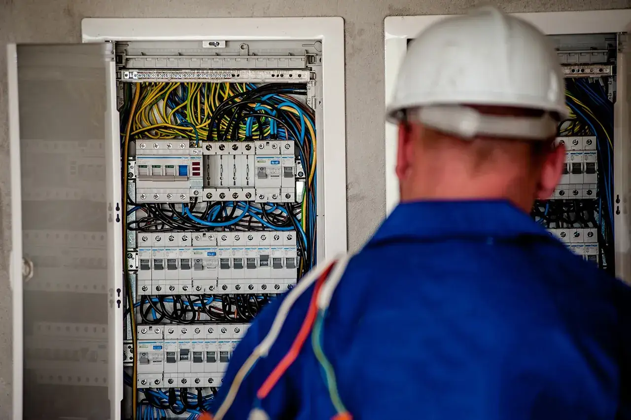 Un tecnico con casco bianco esamina un quadro elettrico pieno di cavi colorati. Un passo fondamentale per capire come diventare elettricista.