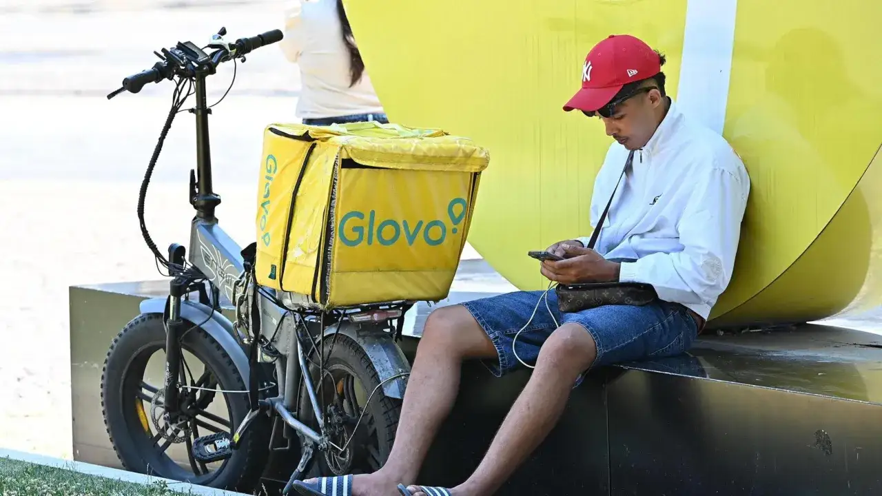 Giovane rider con zaino giallo Glovo controlla il telefono, pronto a lavorare. Bicicletta elettrica parcheggiata.