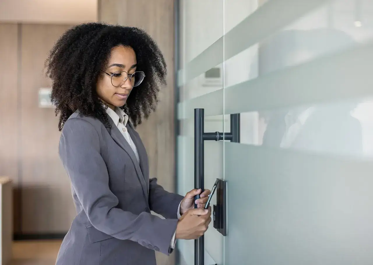 Donna con giacca grigia e occhiali apre una porta di vetro con il telefono, forse per evitare il micro management.