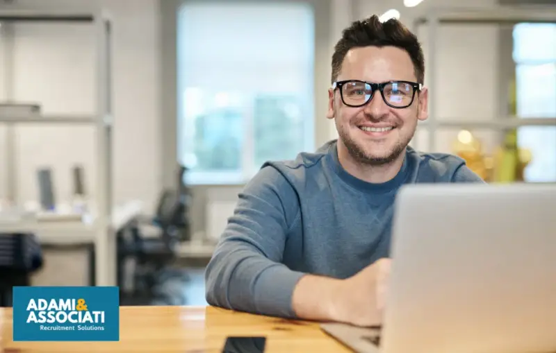Uomo sorridente con occhiali, seduto alla scrivania con un laptop. Sembra un plant manager che gestisce il suo team.